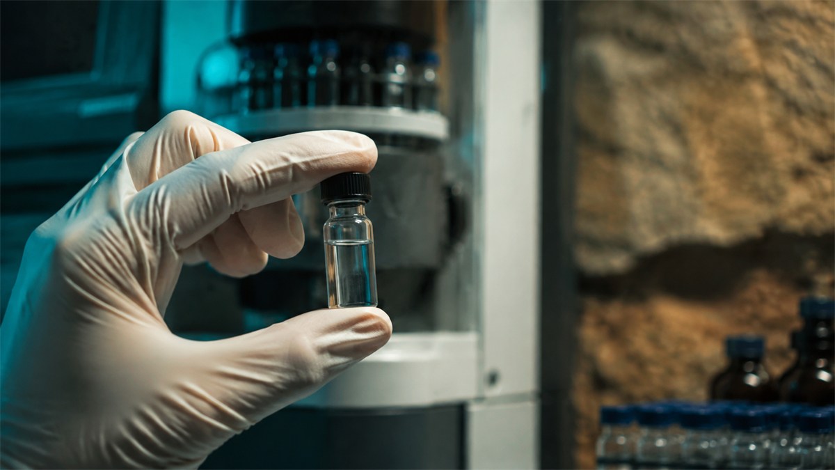 A gloved hand holds a capped vial beside laboratory equipment.